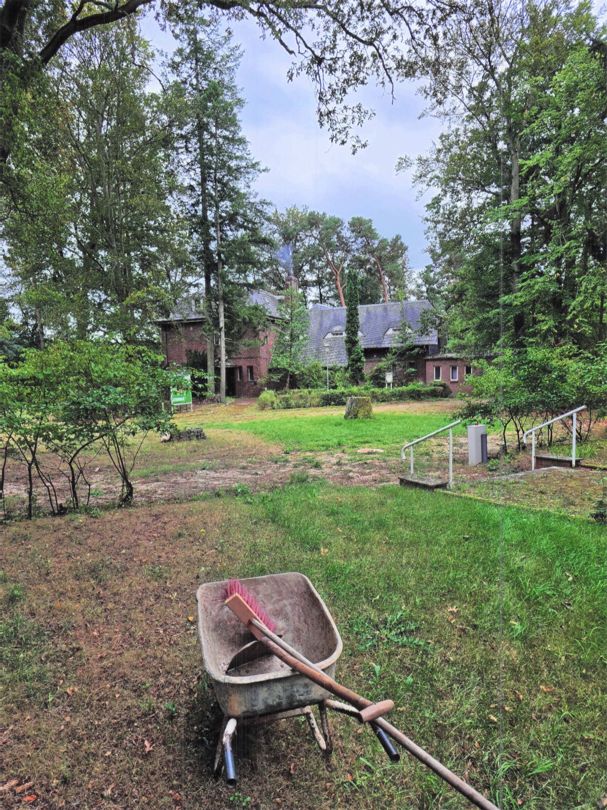 Verwilderter Garten mit alter Schubkarre, im Hintergrund das Waldhotel Seehof